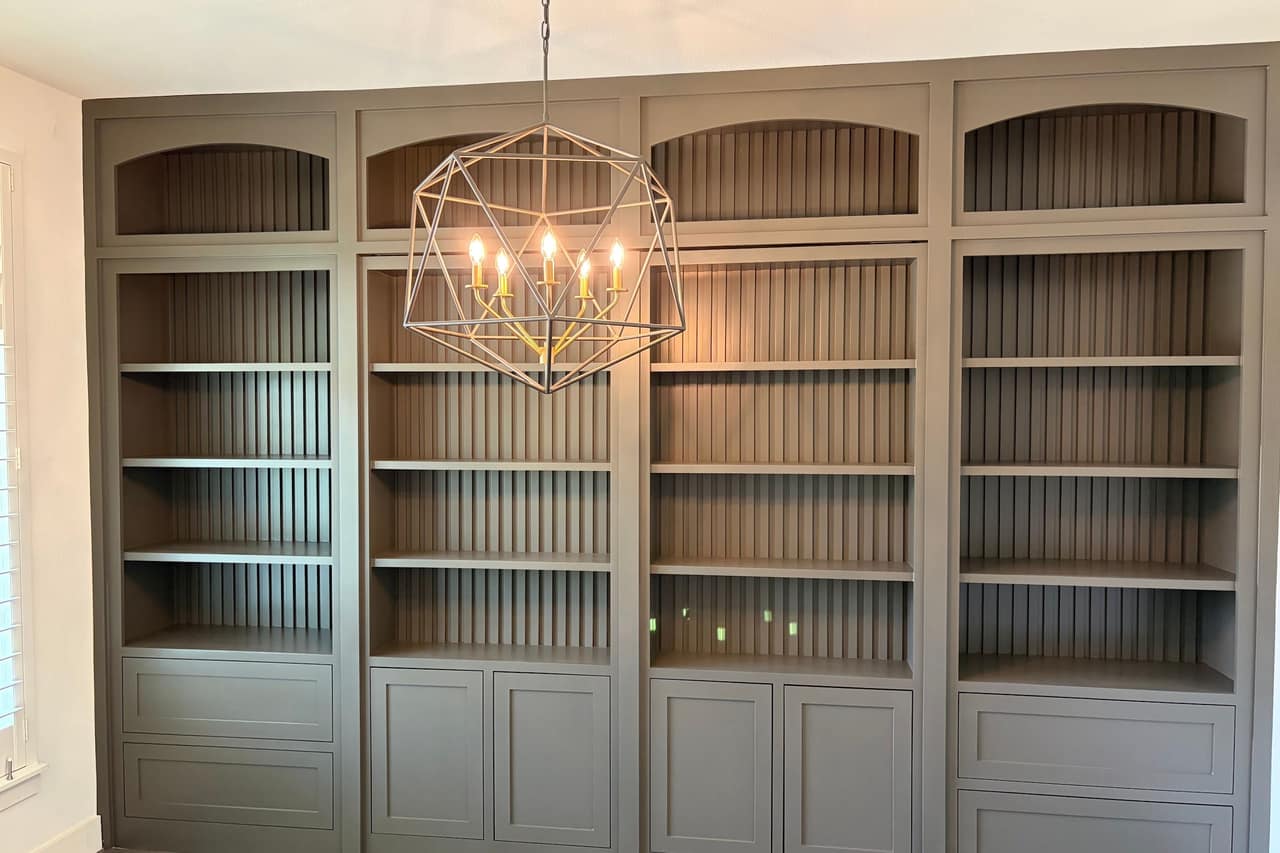 Floor-to-ceiling built-in bookcases painted soft sage green, with beadboard backs and a brass chandelier above